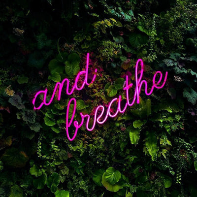 "Glowing pink ‘And Breathe’ LED neon sign mounted on a green foliage wall, adding a relaxing, stylish accent to any room. Perfect for custom neon designs, this sign by London Neon combines vibrant LED lighting with affordability and eco-friendly materials. Visit our store for pre-made and custom LED neon sign options, with a price match guarantee across the UK."